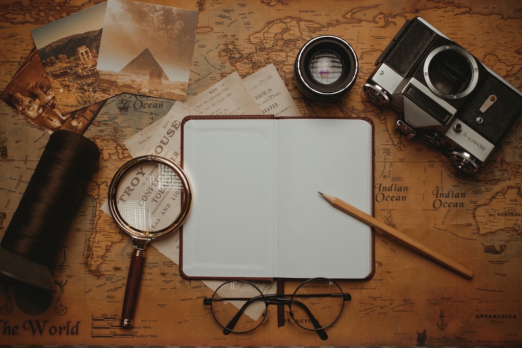magnifying glass, camera and lens, notepad and pencil atop an aged map along with some postcards