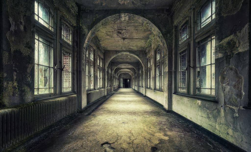 Long, abandoned corridor in a decaying building, with cracked floors, peeling walls, tall windows on both sides, and a series of arches leading towards a dark doorway in the distance.