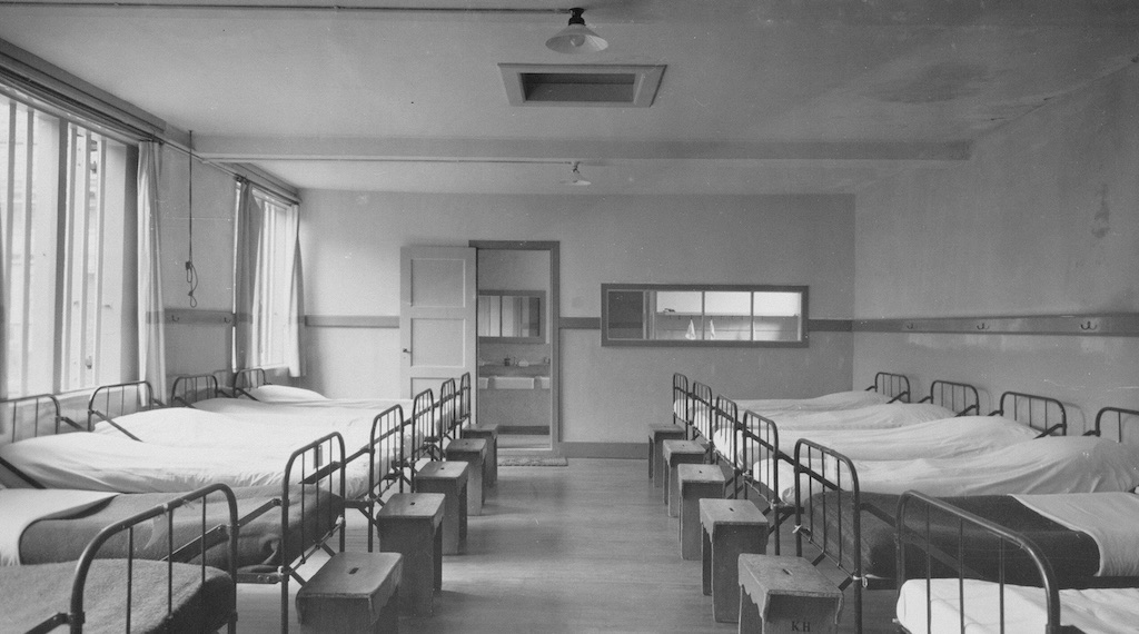 Black-and-white photograph of a large dormitory or hospital ward with rows of metal-framed beds lining both sides of the room, small bedside stools beside each bed, tall windows along the left wall, and a doorway and interior window at the far end.