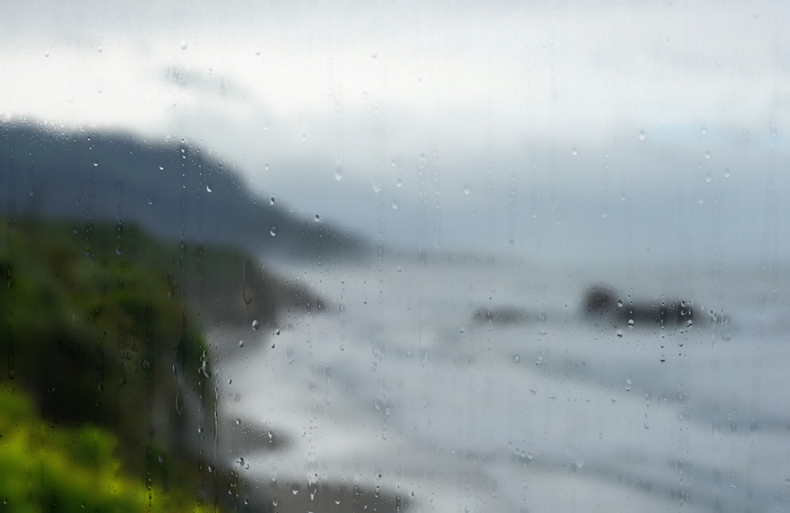 Rain on glass over a soft-focus coastline: green cliffs falling to a grey-blue sea, distant headland, low cloud.