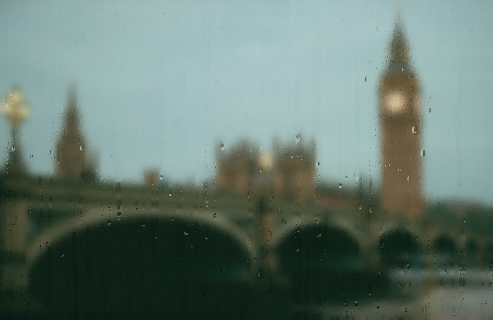 Rain on glass over Westminster Bridge and Big Ben at dusk: a teal-grey London sky, lamps just beginning to glow on the bridge.
